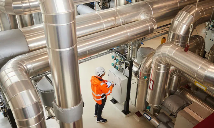Technician in orange safety jacket and hard hat inspecting control panel amid large stainless steel industrial pipes.