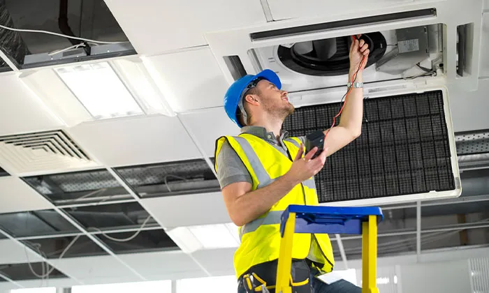 HVAC technician in blue hard hat and hi‑vis vest on ladder testing ceiling air conditioning unit with multimeter