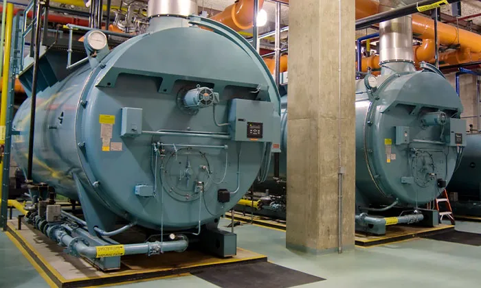 Two teal industrial boilers in a commercial boiler room with piping, valves, and a concrete support column
